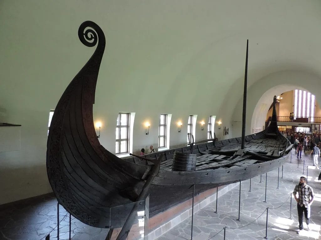 Oseberg ship on display in Oslo: an oak-built longship with high prow and sweeping lines set in a white gallery, showing the kind of vessel that linked Scandinavian harbors with distant coasts.