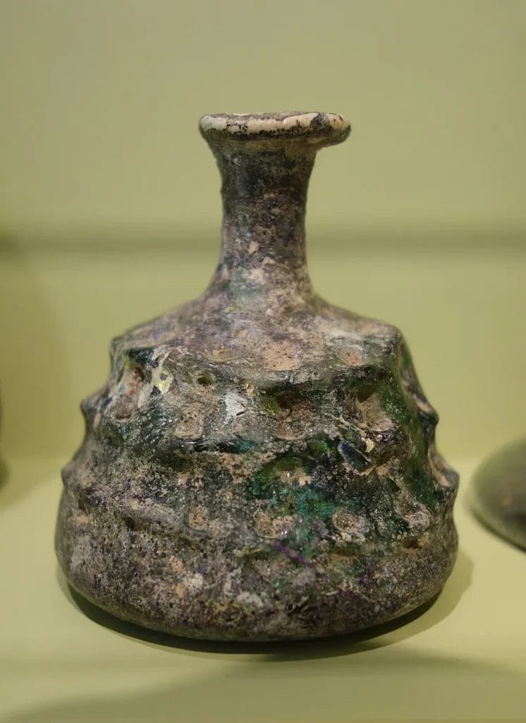 Narrow glass unguentarium with pinched decoration, museum display.